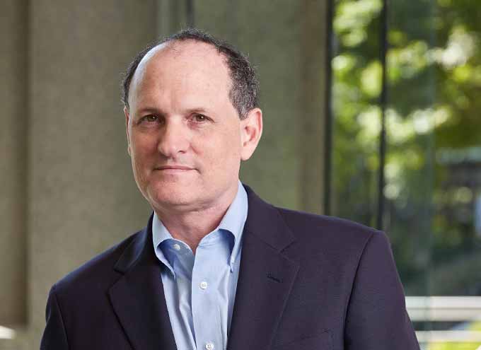 A man in a suit jacket and blue shirt stands indoors near a window with greenery visible outside.